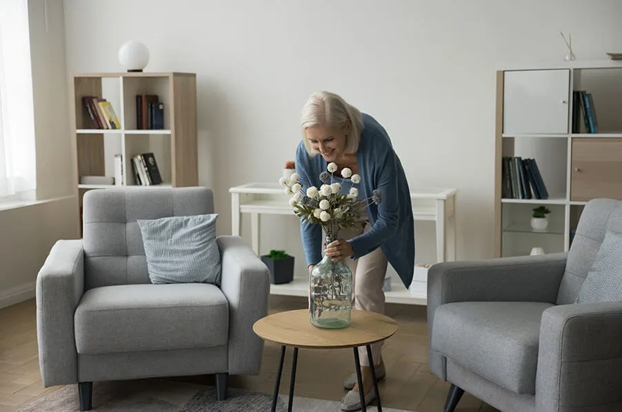 Senior woman placing a vase of flowers on a small round, wooden table.